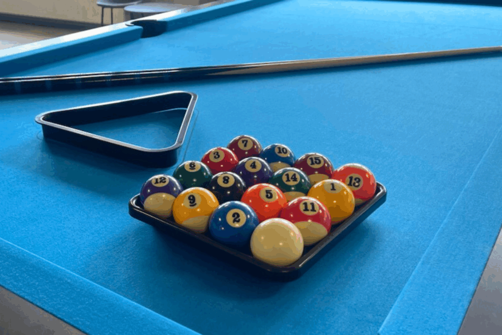 Billard Kugeln sind in einem viereckigen Rahmen plaziert, daneben liegt ein Rahmen in Form eines Dreiecks und zwei Queues. Dies liegt auf einem blauen Billard Tisch
