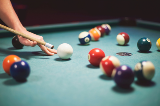 Ein grüner Billard Tisch mit Billard Kugeln in verschiedenen Farben. Eine Person schlägt gerade mit einem Queue an eine weiße Kugeln.