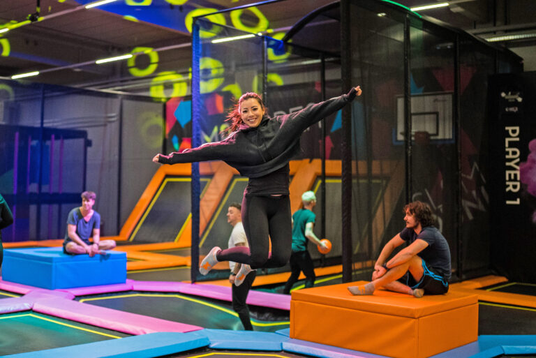 Eine Frau springt in einer Trampolinhalle in dem Indoor Freizeitpark MAXX Arena. Im Hintergrund ist der Trampolinpark zu sehen und vier weitere Personen.