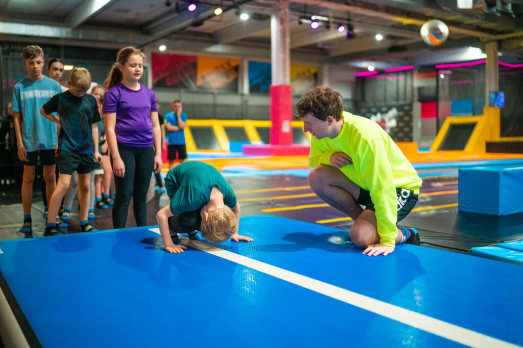 Ein kleiner Junge macht einen Purzelbaum auf einem Trampolin. Dahinter stehen weitere Kinder und ein Trainer in neongelbem Pullover sieht zu.