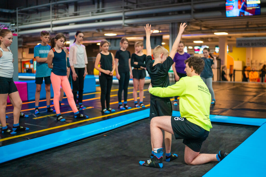 Ein kleiner Junge möchte einen Salto probieren, ein Trainer hilft ihm dabei und hält ihn fest. Im Hintergrund sind weitere kleine Kinder.