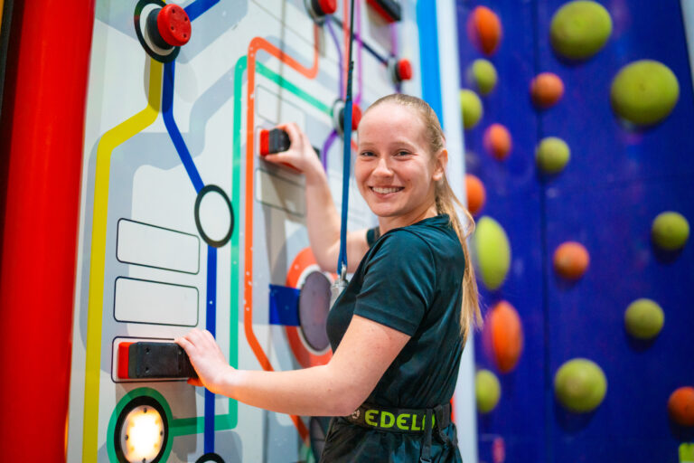 Ein Mädchen klettert eine interaktive Kletterstation, sie ist im Portrait fotografiert. Auf dieser Kletterwand leuchten ebenso gründe Lichter, welche betätigt werden müssen.