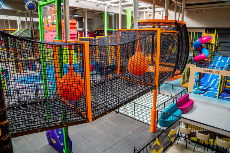 Ein drei Meter hohes Höhenlabyrinth in einer Indoor Halle. Die Umrandung ist orange und das Netz schwarz. In dem Labyrinth sind verschiedene Hindernisse. Im Hintergrund sind blau pinke Rutschen.