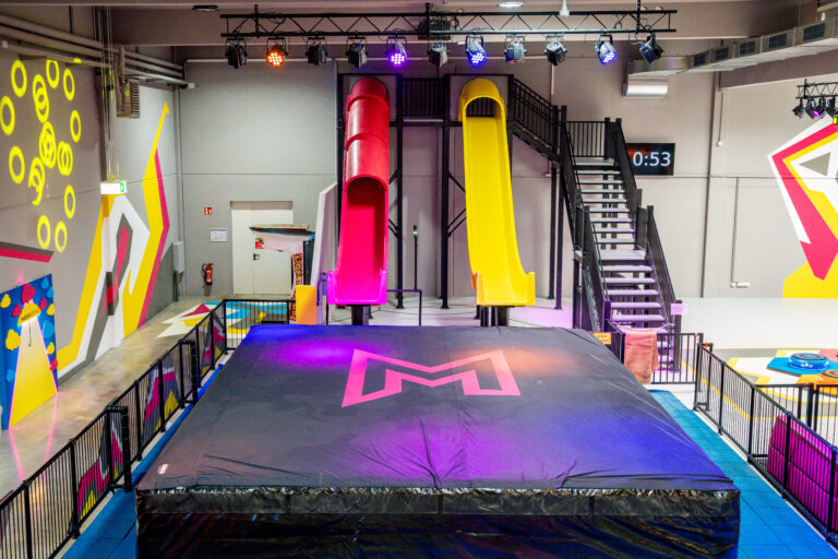 Zwei 6-Meter-Rutschen stehen in gelb und pink nebeneinander. Unter den Rutschen liegt ein großer schwarzer Airbag, in welchen dann auf Matten gerutscht wird. Nebendran ist eine Treppe zu sehen, mit welcher man die Rutschen betritt.