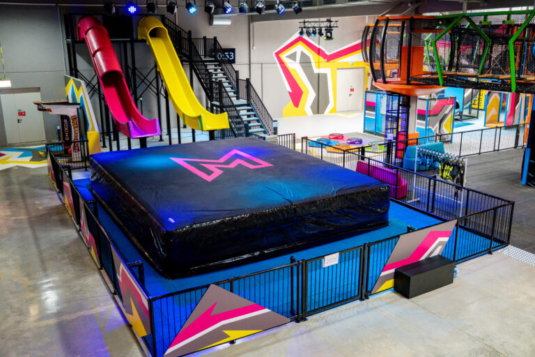 Zwei 6-Meter-Rutschen stehen in gelb und pink nebeneinander. Unter den Rutschen liegt ein großer schwarzer Airbag, in welchen dann auf Matten gerutscht wird. Nebendran ist eine Treppe zu sehen, mit welcher man die Rutschen betritt.