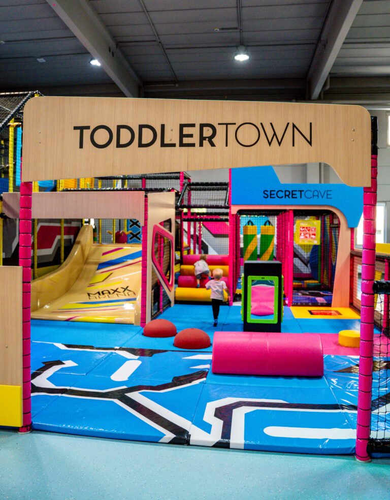 Auf dem braunen Eingangsschild der Area steht Toddler Town. In der Area ist eine weiche blaue Matte als Untergrund aufgelegt. Viele kleine Hindernisse und interaktive Spiele für Kleinkinder sind aufgebaut.