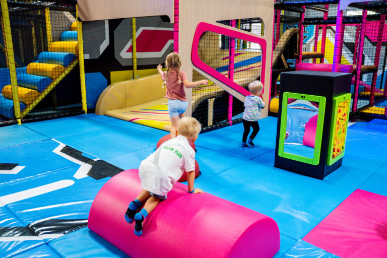 Drei kleine Kinder spielen in der Toddler Town. Als Boden dient eine blaue Schaumstoffmatte, verteilt in der Area sind Spielsachen für Kleinkinder, eine kleine Rutsche sowie kleine Hindernisse. Drei kleine Kinder spielen darin.