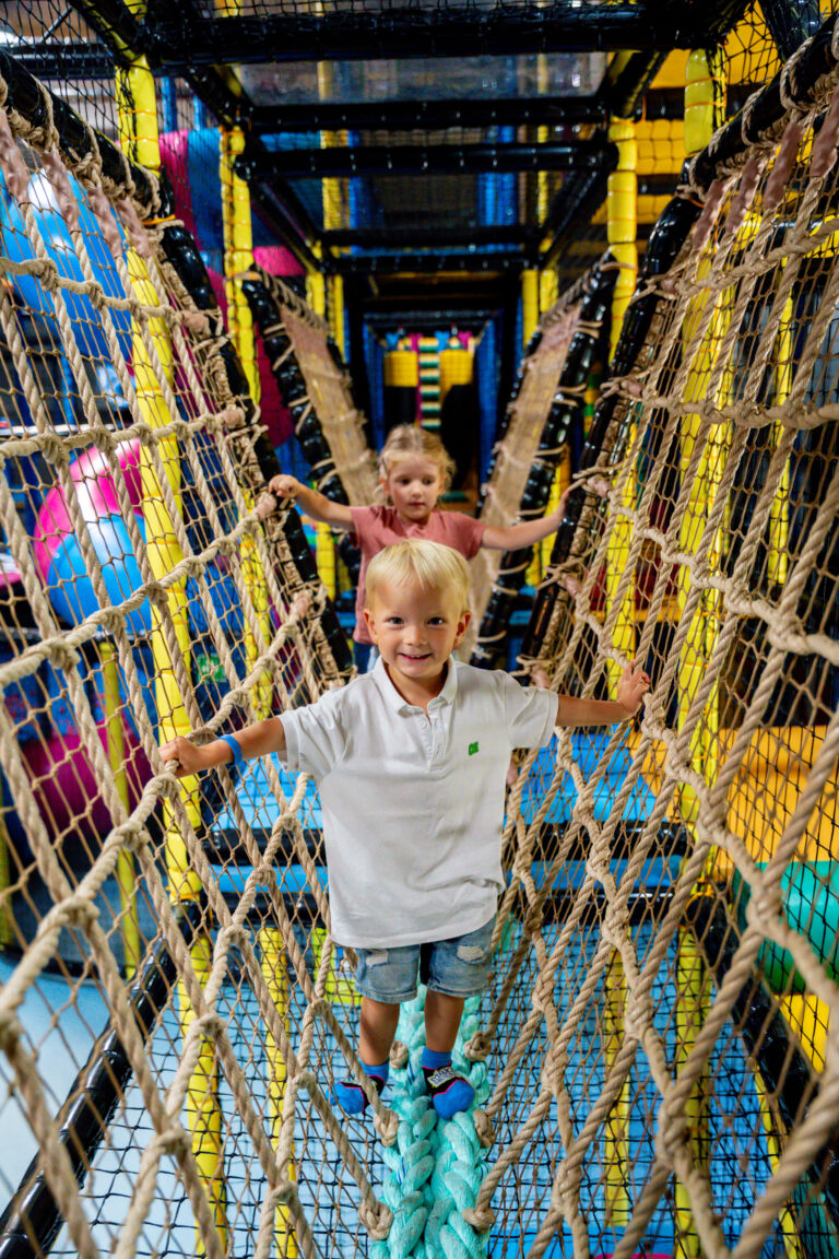 Ein kleiner blonder Junge und ein kleines blondes Mädchen gehen durch ein braunes Netz über einen Balken in einem Indoor Freizeitpark. Beide lachen und halten sich an den Netzen fest.