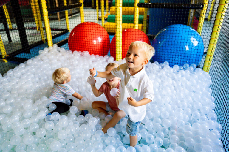 Zwei kleine Jungs und ein kleines Mädchen spielen in einem Bällebad aus durchsichtigen Bällen. Sie liegen in dem Meer voller Bälle und spielen mit diesen. Eingegrenzt sind die Bälle durch ein Netz.