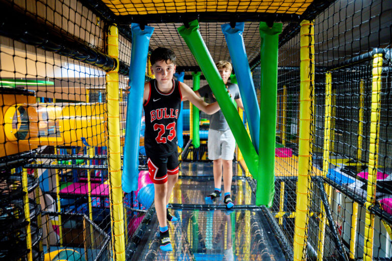 Zwei Jungen in Sportklamotten gehen durch ein mit Netz umrandeten Hindernisbereich in einem Indoor Freizeitpark. Sie gehen durch Hindernisse, gerade durch blaue und grüne Balken.