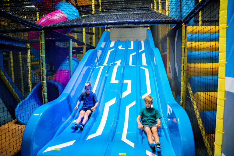 Zwei Jungs rutschen eine blaue weiße Rutsche herunter. Diese hat vier Bahnen nebeneinander, die beiden rutschen auf den zwei äußersten Bahnen.