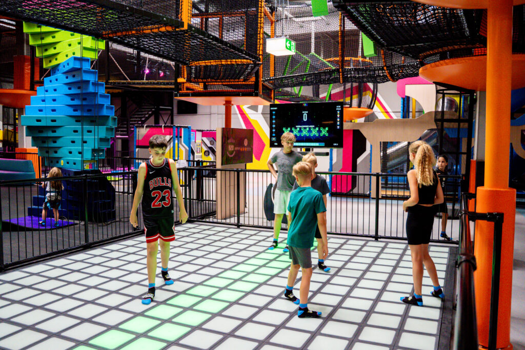 Kinder in Sportklamotten spielen Pixel Games. Sie stehen auf einer Fläche, auf welcher die bunten Pixel Lichter leuchten. Auf einem Bildschirm vor der Fläche wird das Spiel angezeigt. Im Hintergrund ist die Indoor Freizeithalle zu sehen.
