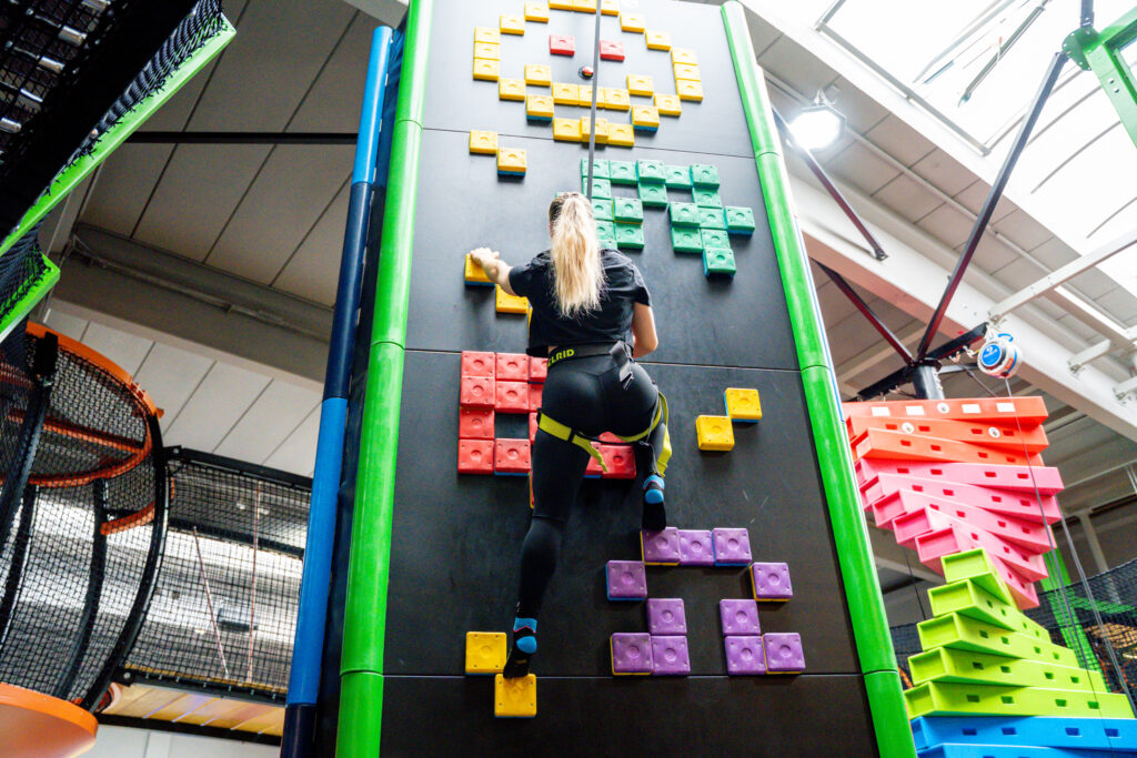 Ein Mädchen klettert eine Kletterwand nach oben, welche schwarz ist und mit bunten Quadraten bestückt ist.