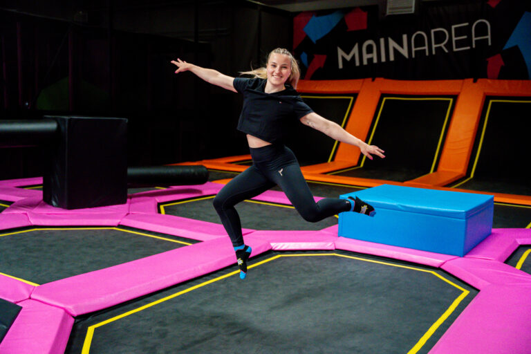 Ein braunhaariges jungendliches Mädchen springt auf einer Trampolinfläche. Sie hat die Arme in der Luft und hat Spaß.
