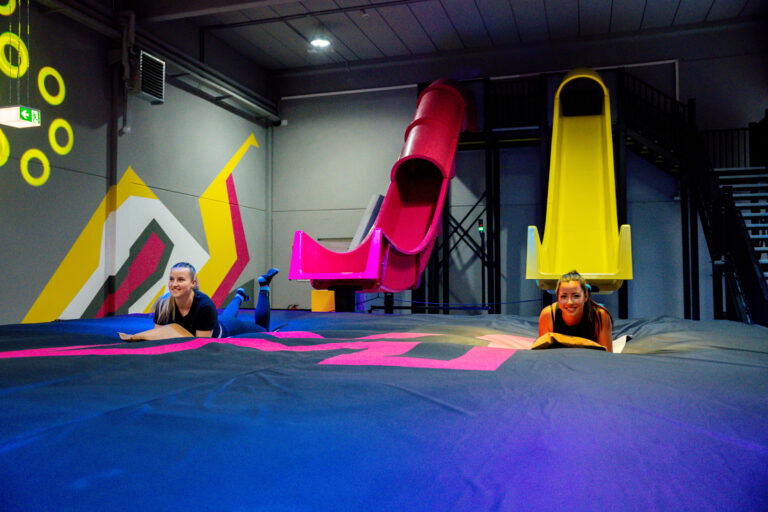 Zwei 6-Meter-Rutschen stehen in gelb und pink nebeneinander. Unter den Rutschen liegt ein großer schwarzer Airbag, in welchen dann auf Matten gerutscht wird. Nebendran ist eine Treppe zu sehen, mit welcher man die Rutschen betritt. Zwei Frauen liegen auf dem Airbag.