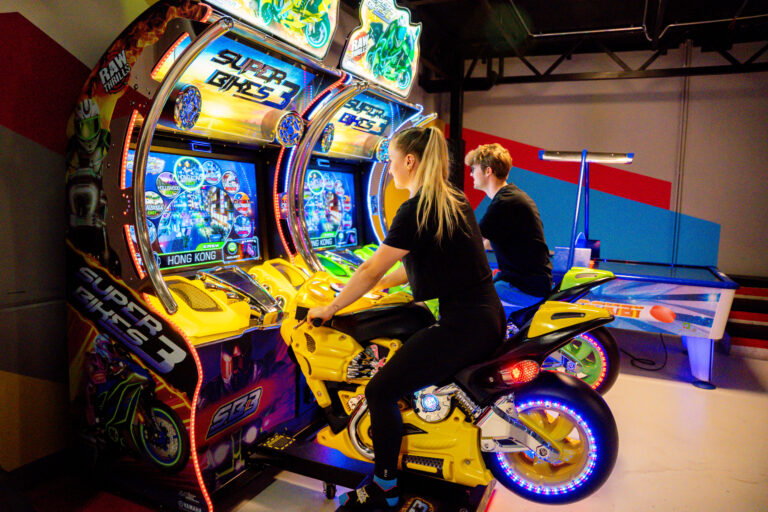 Zwei Jugendliche sitzen auf Motorrädern, welche an einem Bildschirm festgemacht sind und zu einem Videospiel gehören. Sie schauen auf den Bildschirm vor den Motorrädern.