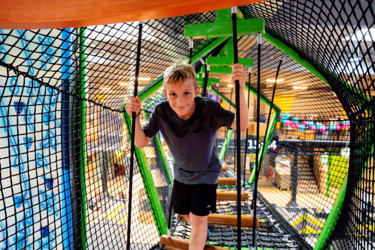 Ein Junge klettert durch ein Höhenlabyrinth und hält sich dabei an dem schwarzen Netz fest, um den Boden nicht zu berühren, sondern lediglich an braunen Holzzylinder zu balancieren.