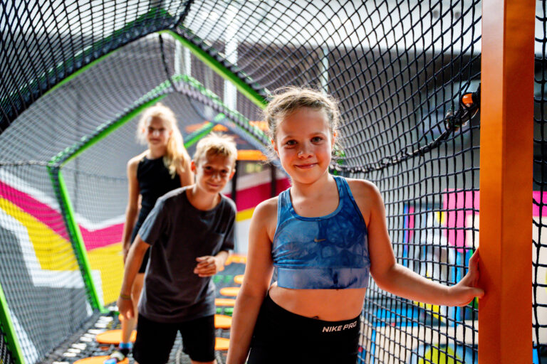 Ein Mädchen in Sportklamotten grinst in die Kamera, im verschwommenen Hintergrund stehen zwei weitere Kinder. Die drei stehen in 3 Meter Höhe in einem Höhenlabyrinth und gehen durch Hindernisse.