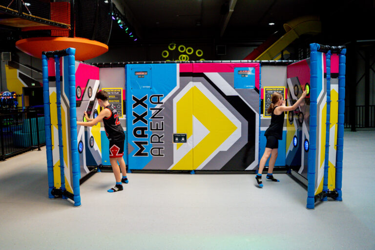 Ein Mädchen und ein Junge stehen in der Skypods Area. Sie spielen gegenüber an der Wand und müssen leuchtende Lichter anklicken. Im Hintergrund ist die Indoorhalle.