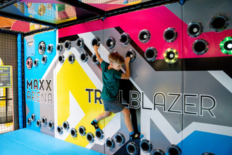 An einer bunten Wand sind Löcher eingearbeitet, welche in verschiedenen Farben Leuchten. An den Löchern kann man sich hangeln. Dies macht ein kleiner Junge in Sportklamotte.