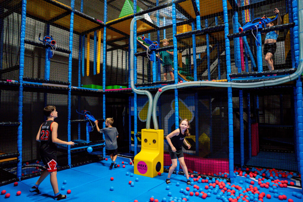 In einer kleinen Arena, welche mit einem Netz umspannt ist, spielen fünf Kinder. Diese müssen sich mit Schaumstoffbällen abwerfen. Die Bälle sind rot sowie blau.