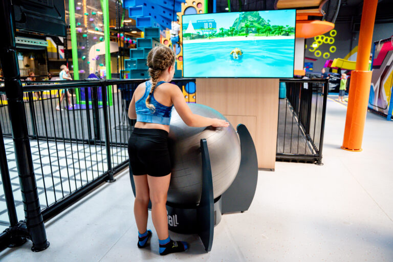 Im Hintergrund ist ein Bildschirm zu sehen, auf welchem eine Videospielfigur abgebildet ist. Im Vordergrund steht ein Mädchen mit einem grauen Ball in der Hand. Dieser Ball ist mit dem Videospiel verknüpft und kann die Videospielfigur steuern.