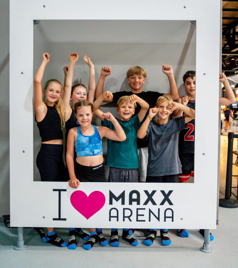Kleine Mädchen und Jungen lachen in die Kamera und heben die Arme in die Luft. Sie stehen in einem weißen Rahmen und vorne steht I love MAXX Arena.