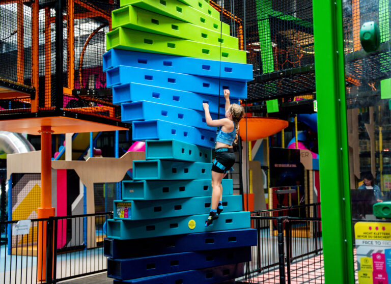 Ein Mädchen klettert eine Kletterstation gesichert nach oben, welche Twister genannt wird und blau ist.