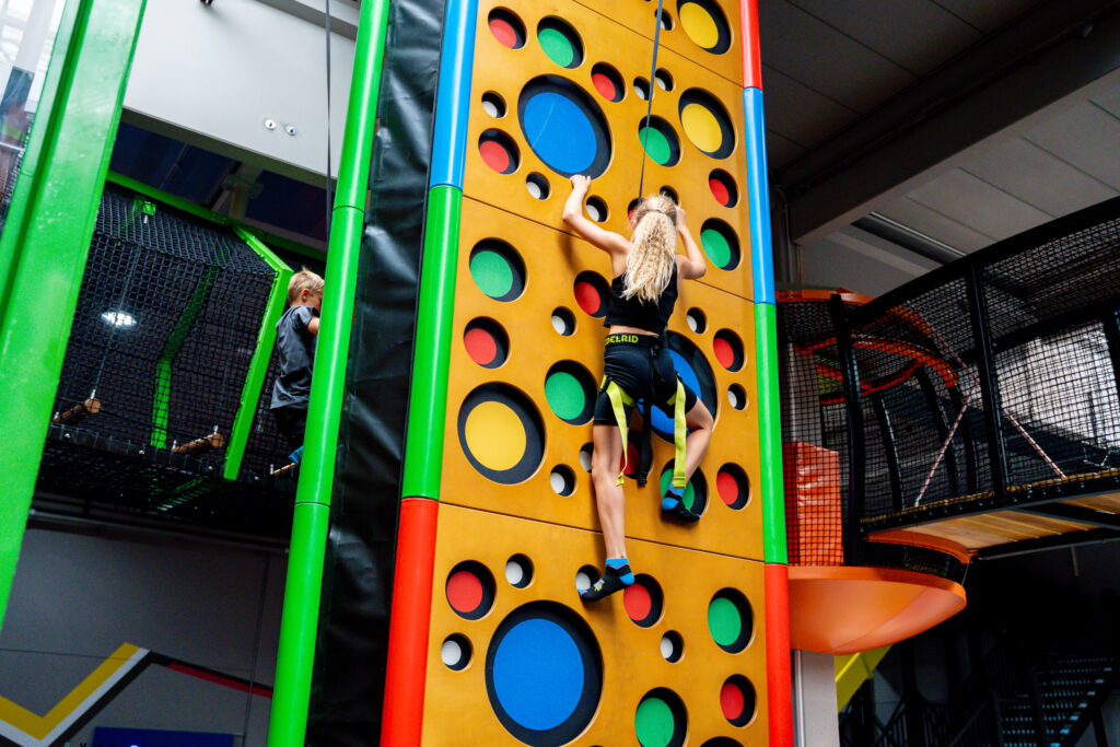 Ein blondes Mädchen klettert gesichert eine Kletterwand nach oben. In der Wand sind bunte Löcher hinterlegt, an welchen sie sich festhalen muss.