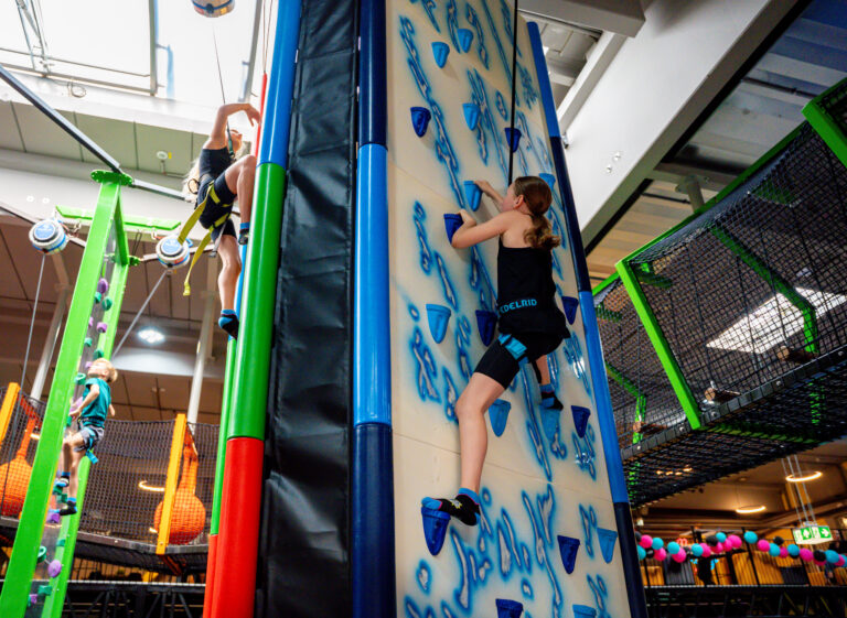 Eine Kletterwand welche weiß ist, jedoch mit blau farbigen Steinen versehen ist, an welchen man nach oben klettern kann. Diese sind als dunkelblaue Eiszapfen dargestellt. Es klettern zwei Mädchen die Wände nach oben.