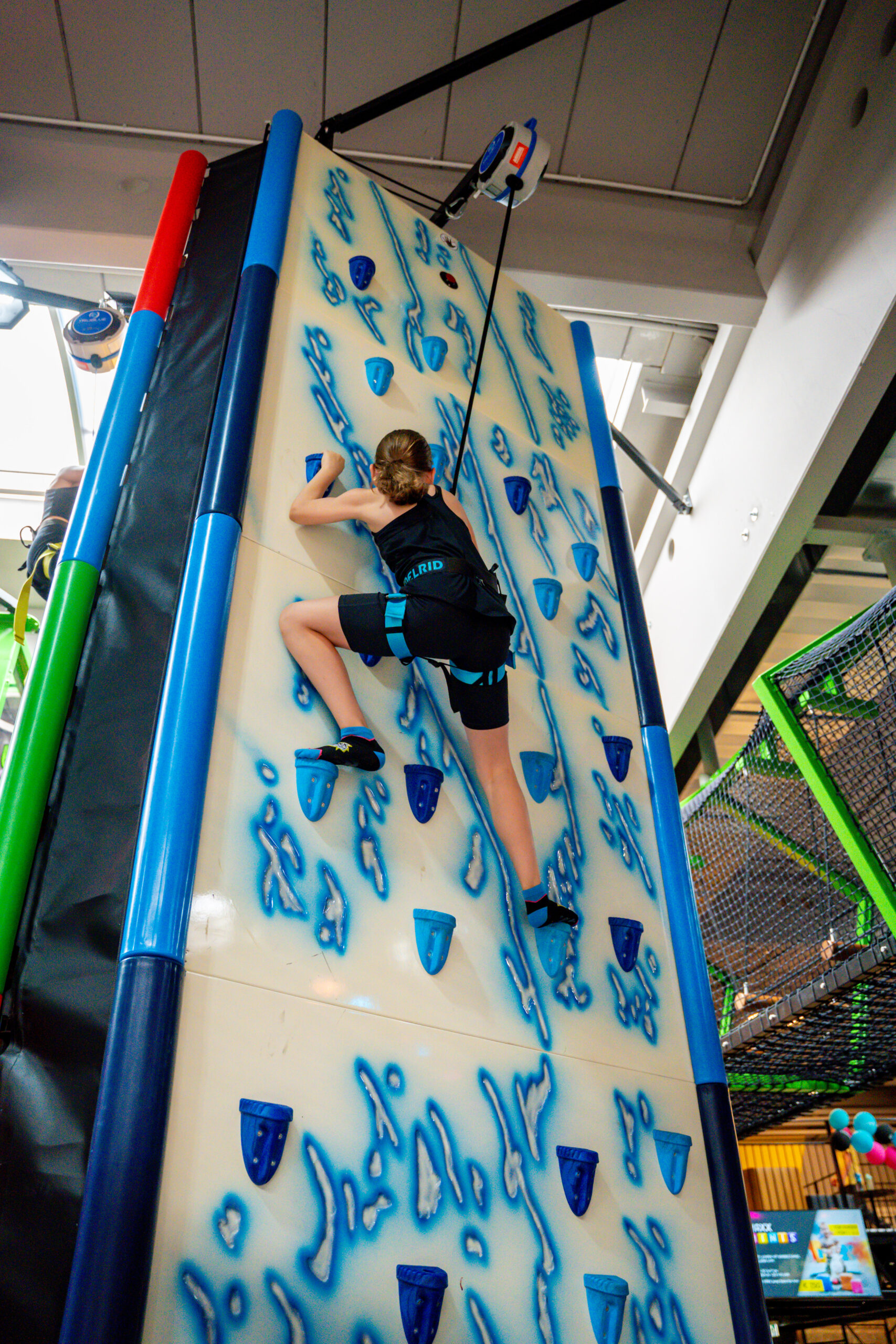 Ein Mädchen klettert eine Kletterwand nach oben, welche weiß ist, jedoch mit blau farbigen Steinen versehen ist, an welchen man nach oben klettern kann. Diese sind als dunkelblaue Eiszapfen dargestellt.