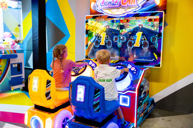 in Spielautomat mit zwei Lenkrädern, bei welchem vorne ein Bildschirm ist. Zwei kleine Kleinkinder sitzen auf den Stühlen und lenken die Autos auf dem Bildschirm.