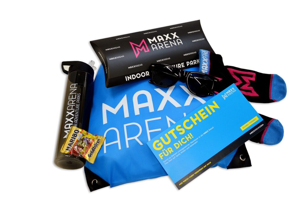 Geschenkartikel sind abgebildet: MAXX Socks, eine kleine Haribo Gummibärchen-Tüte, Trinkflasche in grau, eine Sonnenbrille in schwarz, MAXX Arena Wertgutschein-Karte sowie eine Gutschein-Box.