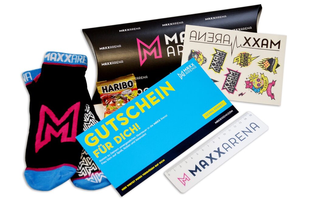 Geschenkartikel sind abgebildet: MAXX Socks, Lineal mit MAXX Arena Logo, eine kleine Haribo Gummibärchen-Tüte, Klebe-Tattoos, MAXX Arena Wertgutschein-Karte sowie eine Gutschein-Box.
