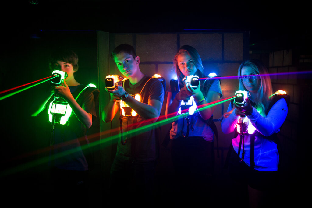 Vier Erwachsene in einer dunklen Arena tragen leuchtende Lasertag-Westen und phasern lachend mit ihren Phasern in die Kamera. Bunte Laserstrahlen in Grün und Rot sind sichtbar.