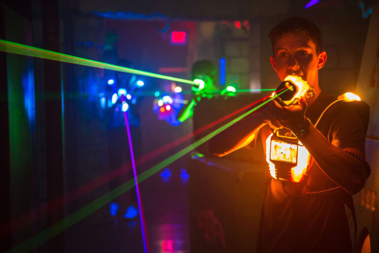 Ein Jugendlicher mit leuchtender Lasertag-Weste phaserd mit seinem Phaser nach vorne. Im Hintergrund sind weitere Personen mit bunten Licht auf der Weste sichtbar.