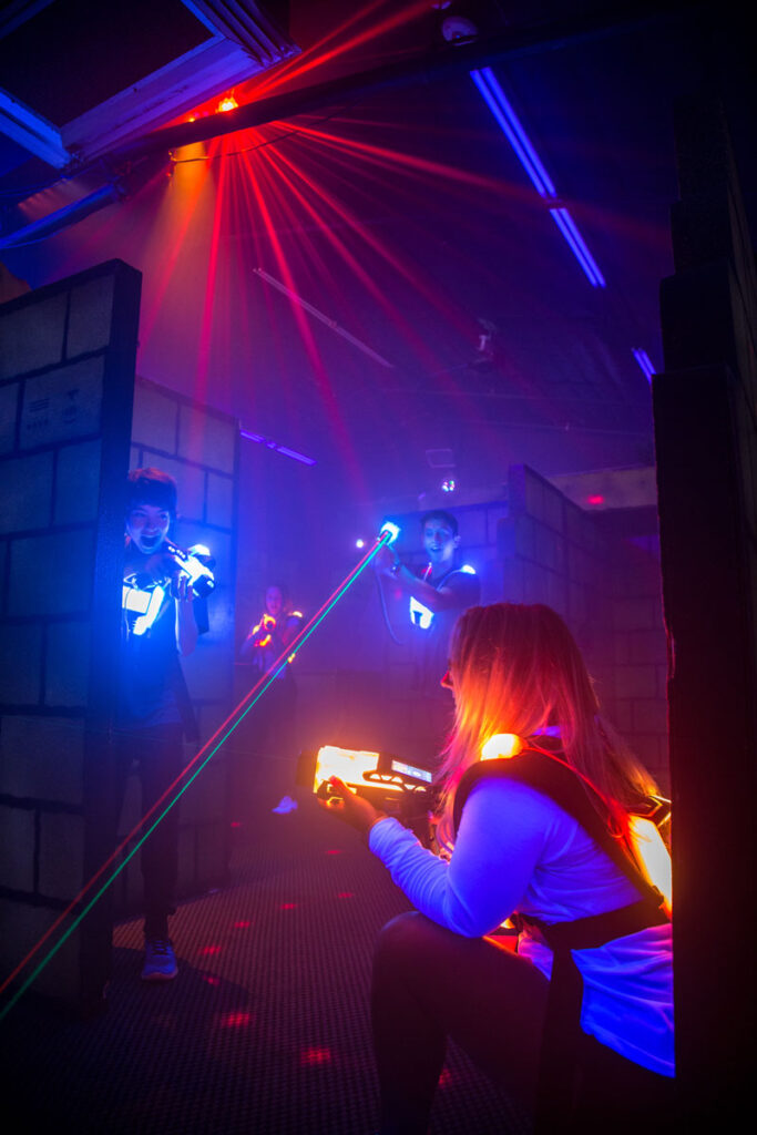 Eine junge Frau mit langen blonden Haaren und Brille lacht und markiert mit einem Lasertag Phaser. Um sie herum sind mehrere Mitspieler mit leuchtenden Westen zu sehen. Bunte Laserstrahlen und Lichtstrahlen in Rot, Grün und Blau durchziehen die Arena.