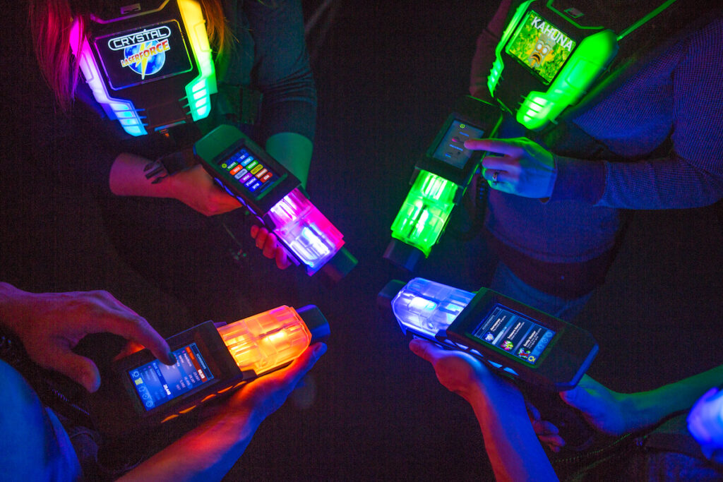 Nahaufnahme mehrerer Lasertag-Westen und -Phasern mit bunten, leuchtenden Farben in Grün, Blau, Rot und Orange. Mehrere Hände halten die Geräte, die Displays und Knöpfe zeigen.