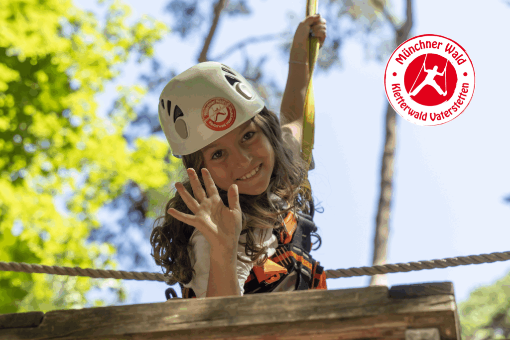 Ein junges Mädchen mit Helm liegt auf einer Plattform eines Parcours im Kletterwald, Münchner Wald. Sie winkt und lächelt in die Kamera.