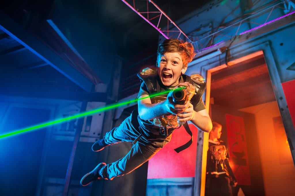 Ein Junge in einer Lasertag-Weste springt lachend in die Luft und zielt mit einem Lasertag Phaser direkt nach vorne. Ein grüner Laserstrahl ist sichtbar, die Arena ist bunt mit blauem und orangem Licht beleuchtet. Im Hintergrund steht eine weitere Person in einer ähnlichen Weste.