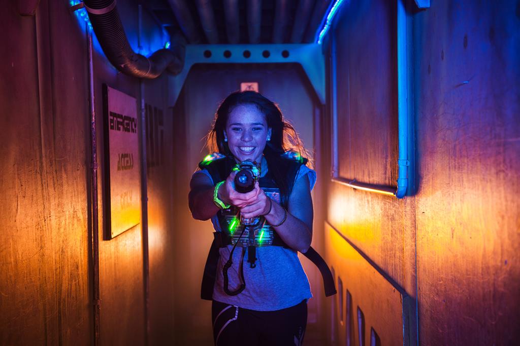 Eine junge Frau mit braunen Haaren lächelt und hält einen Lasertag Phaser in der Hand. Sie trägt eine leuchtende Lasertag-Weste in einer dunklen Arena mit bunten Lichtern in Blau und Orange.