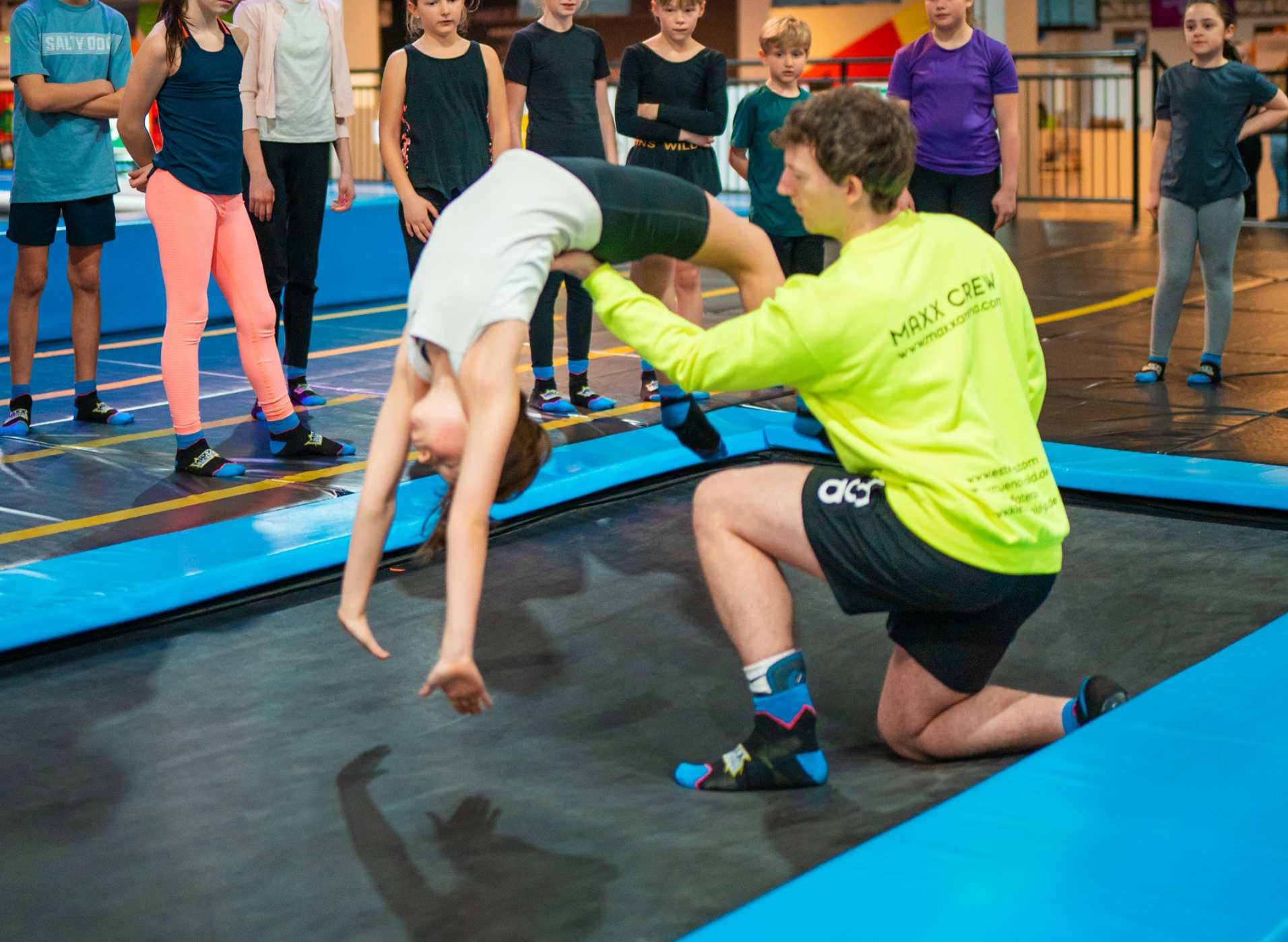 Es ist ein Feriencamp Trainer der MAXX Arena zusehen, welcher einem Mädchen zeigt wie sie ihre Arme für einen Rückwärts Flick-Flack halten sollte.