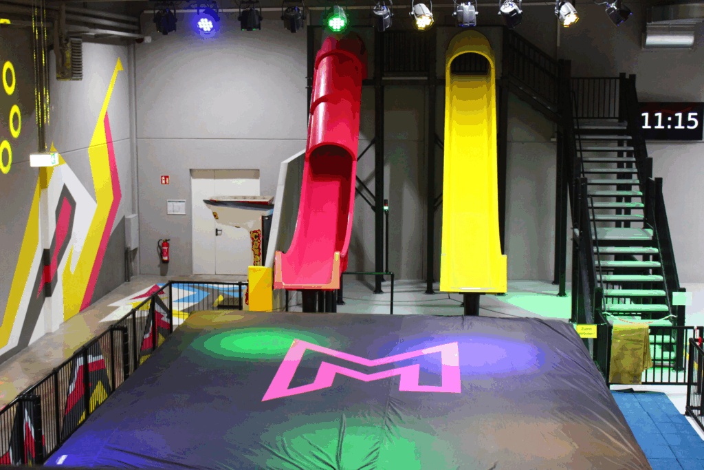 Zwei 6-Meter-Rutschen stehen in gelb und pink nebeneinander. Unter den Rutschen liegt ein großer schwarzer Airbag, in welchen dann auf Matten gerutscht wird. Nebendran ist eine Treppe zu sehen, mit welcher man die Rutschen betritt.