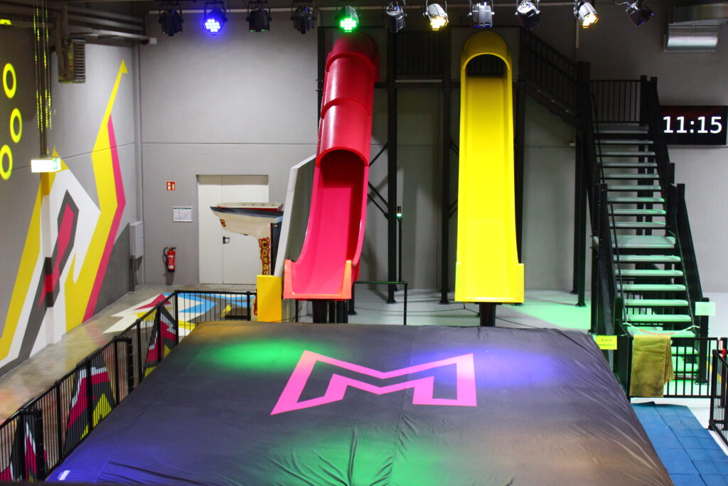 Zwei 6-Meter-Rutschen stehen in gelb und pink nebeneinander. Unter den Rutschen liegt ein großer schwarzer Airbag, in welchen dann auf Matten gerutscht wird. Nebendran ist eine Treppe zu sehen, mit welcher man die Rutschen betritt.
