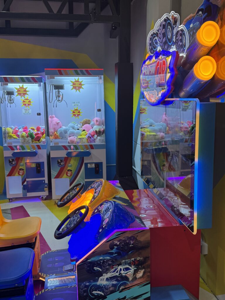 Ein Spielautomat mit zwei Lenkrädern, bei welchem vorne ein Bildschirm ist. Im Hintergrund sind Spielautomaten, aus welchem man mit einem kleinen Kran Stofftiere fischen kann.