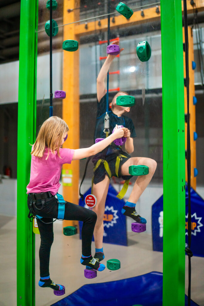 Zwei KInder, ein Junge und ein Mädchen, machen eine Kletterchallenge mit einer Kletterstations Face2Face. Dabei Klettern sie gegenüber in die Höhe und sehen sich durch eine Plastikscheibe.