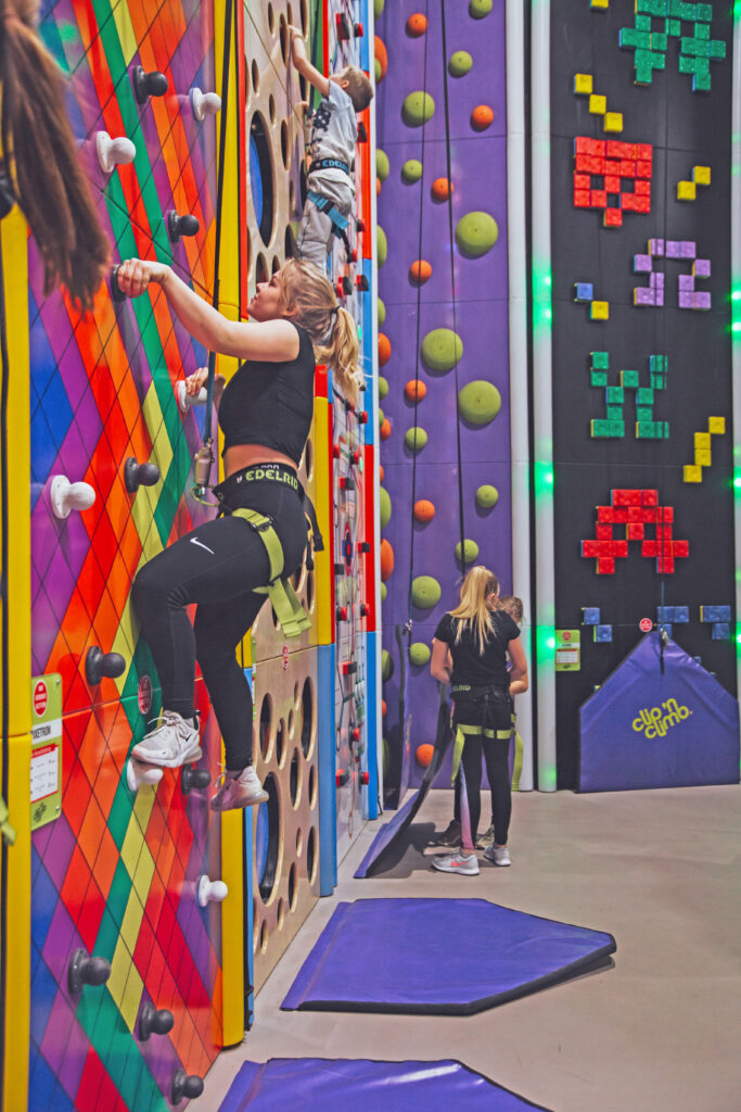 Ein Mädchen klettert in die Höhe, auf der Kletterwand sind hell gefärbte Pixel in unterschiedlichen Farben zu sehen. Im Hintergrund klettern drei weitere kleine Kinder.