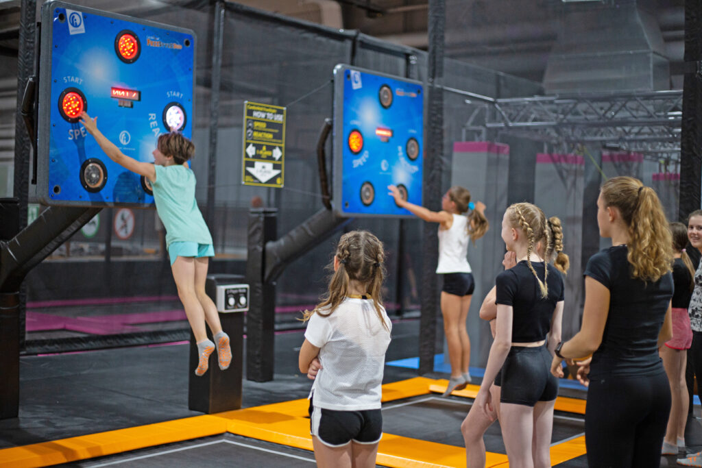 Mehrere Kinder stehen in einer Schlange um das Spiel Cardio Wall zu spielen. Zwei Mädchen spielen dies bereits und hüpfen auf jeweils einem kleinen Trampolin in die Höhe, um schnell aufleuchtende Buttons so schnell wie möglich zu berühren.