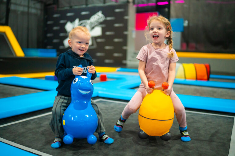 Zwei Minis, links ein Junge und rechts ein Mädchen, sitzen lachend auf Gummitieren und hüpfen währenddessen auf einem Trampolin für Babys.