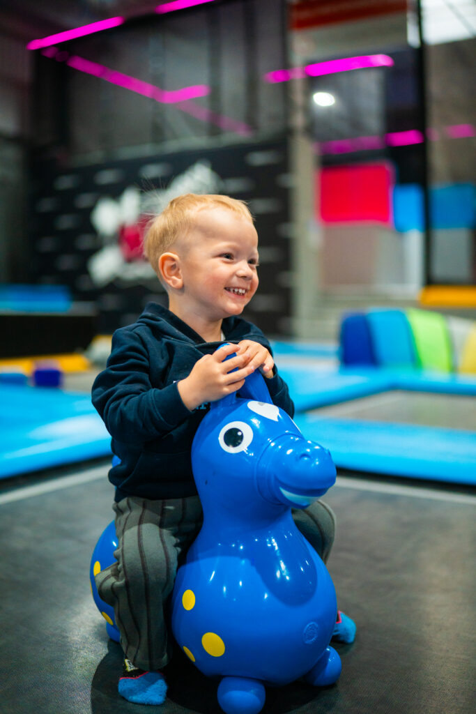 Ein Junge sitzt lachend auf einem baluen Gummi-Pferd und hüpft währenddessen auf einem indoor Trampolin für Kleinkinder.